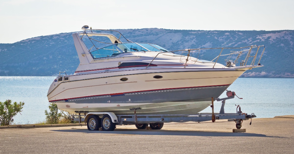 White cabin cruiser on a dual-axle trailer beside calm water with distant hills beneath a clear blue sky.