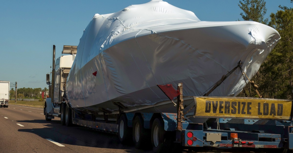 Shrink-wrapped yacht secured on an oversized transport trailer traveling along a highway under clear blue sky.