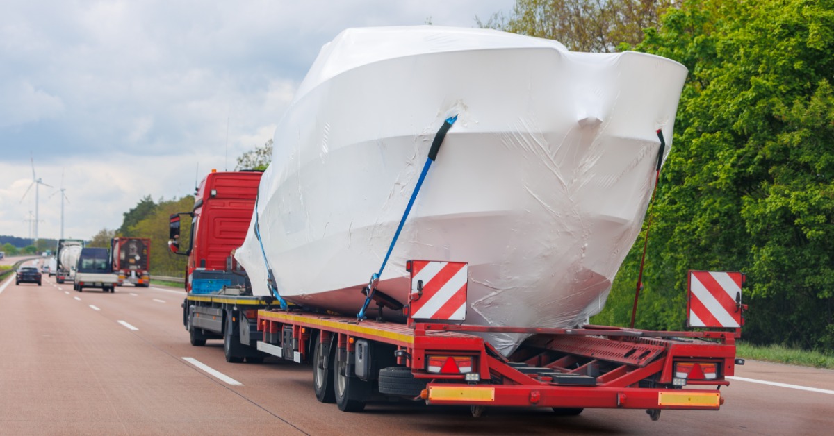 Shrink-wrapped boat on a red flatbed trailer traveling along a highway beside green trees under cloudy skies.