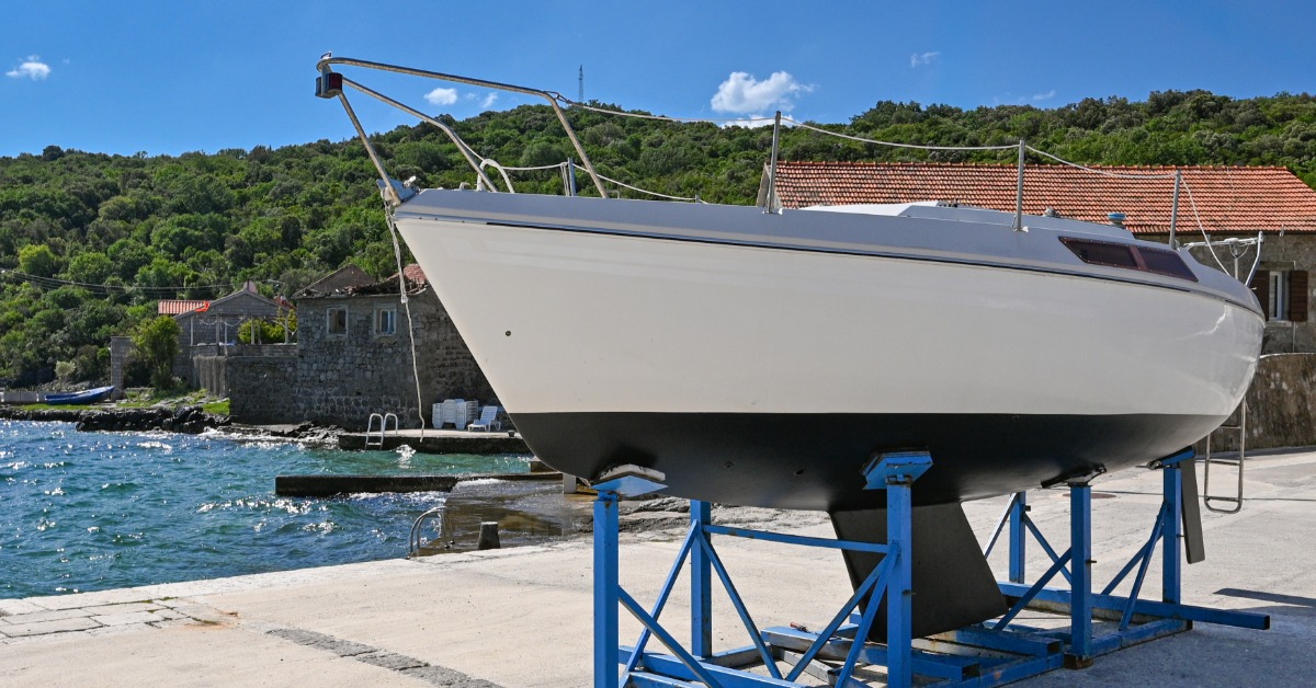 Crane lifts a luxury sailboat above a transport truck in a boatyard as workers prepare it for overland shipping.