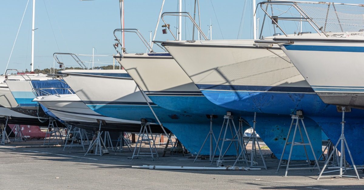 Row of sailboats resting on metal stands in a boatyard, with blue hulls and tall masts stretching into a clear sky.