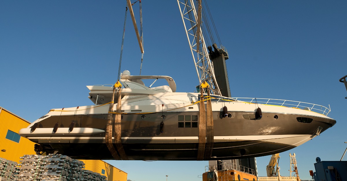 Large luxury yacht hangs from crane straps above an industrial shipyard, with stacked materials and cranes nearby.