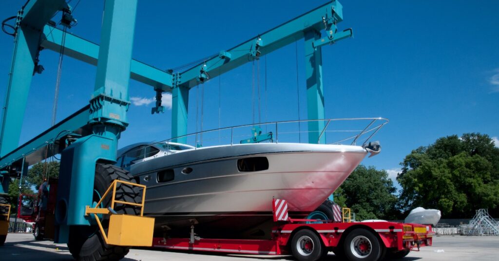Large silver yacht rests on a red transport trailer beneath towering blue marina cranes on a clear, sunny day.
