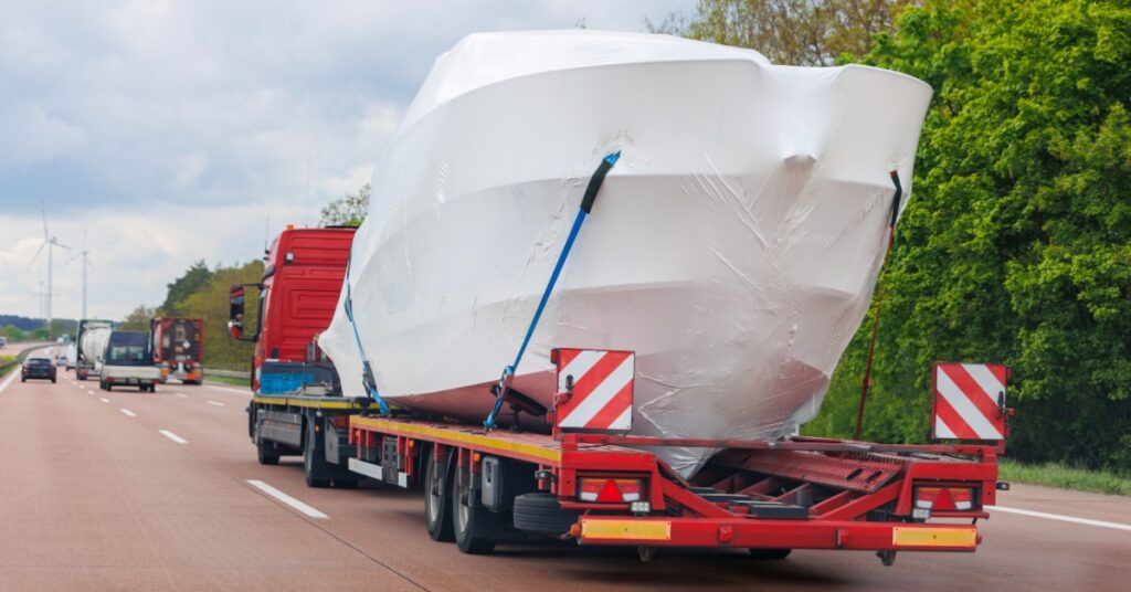 Large yacht wrapped in white protective covering is secured to a red transport trailer traveling on a highway.