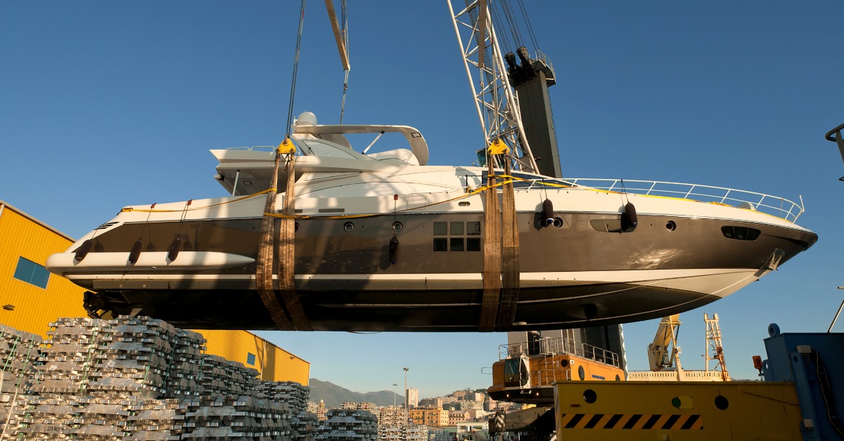 Large luxury yacht suspended by crane straps above an industrial dock, with stacked metal cargo and port equipment below.