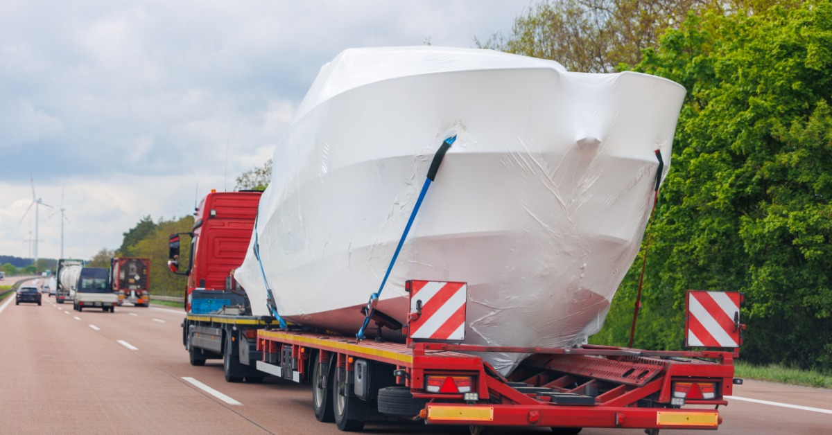 Large shrink-wrapped yacht secured to a red flatbed trailer traveling on a highway bordered by green trees.