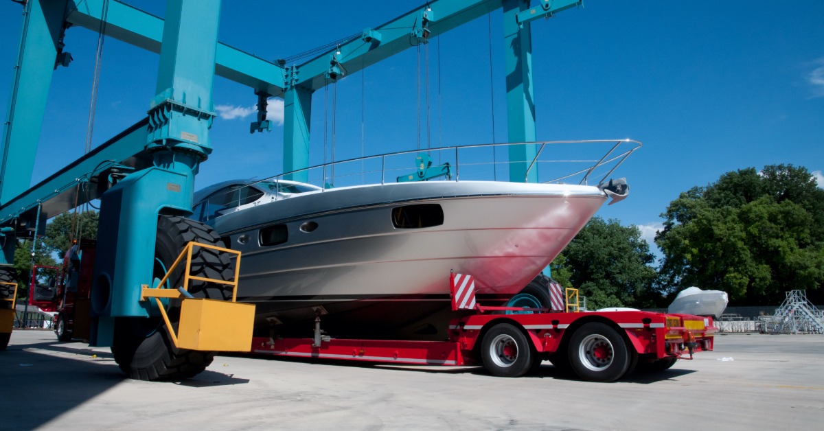 Large white yacht positioned on a red transport trailer beneath towering blue boat lifts at an outdoor marina yard.