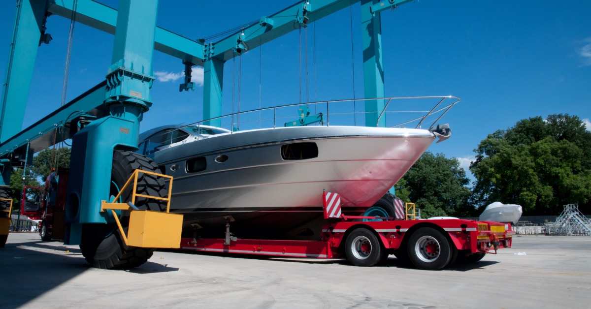 Large white yacht rests on a red transport trailer beneath towering blue boat-lift cranes at a marina.