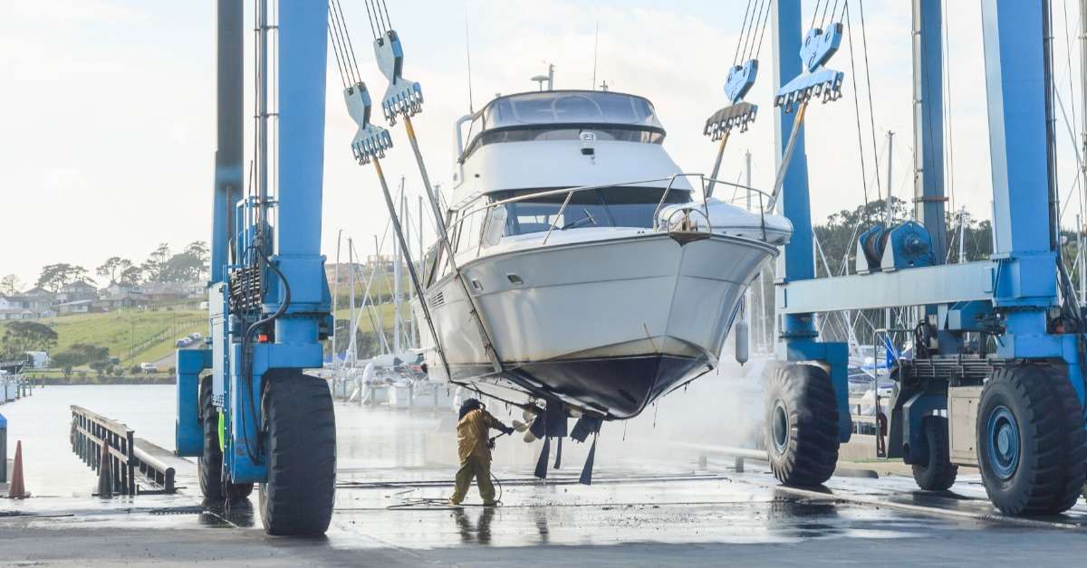 Large white yacht hangs between two blue boat lifts as a worker cleans its hull at a marina beside the water.
