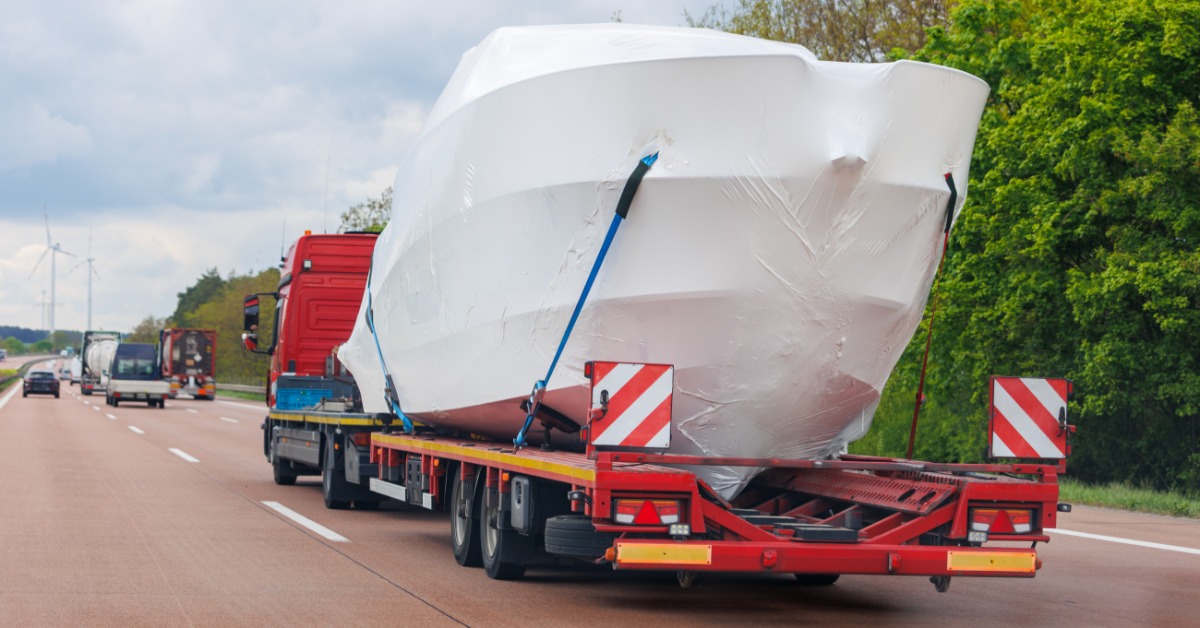 Large yacht wrapped in white protective covering is secured to a red transport trailer traveling on a highway.