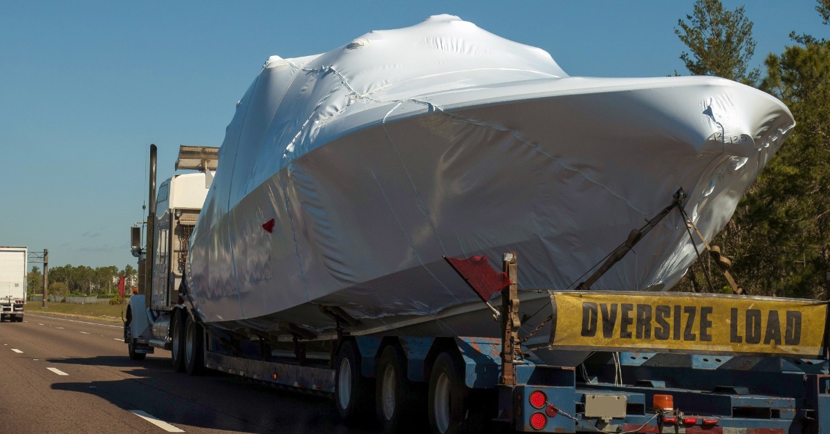 White-wrapped yacht secured on a transport trailer traveling down a highway with a yellow oversize load banner.