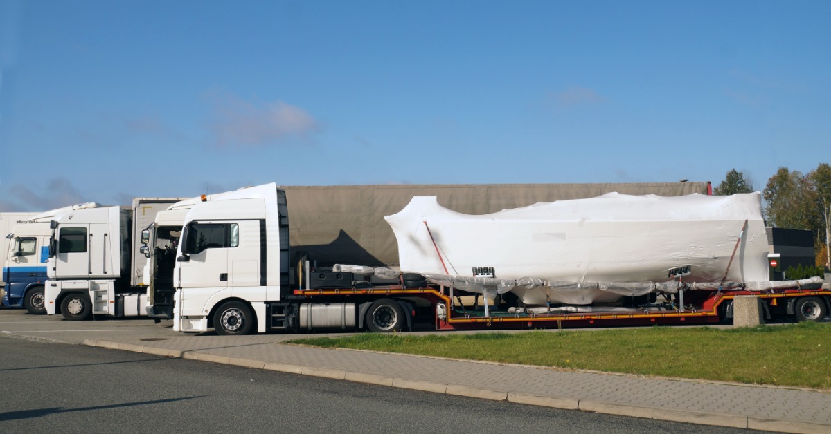 White semi-truck hauls a large yacht wrapped in protective covering on a long flatbed trailer beside a roadway.