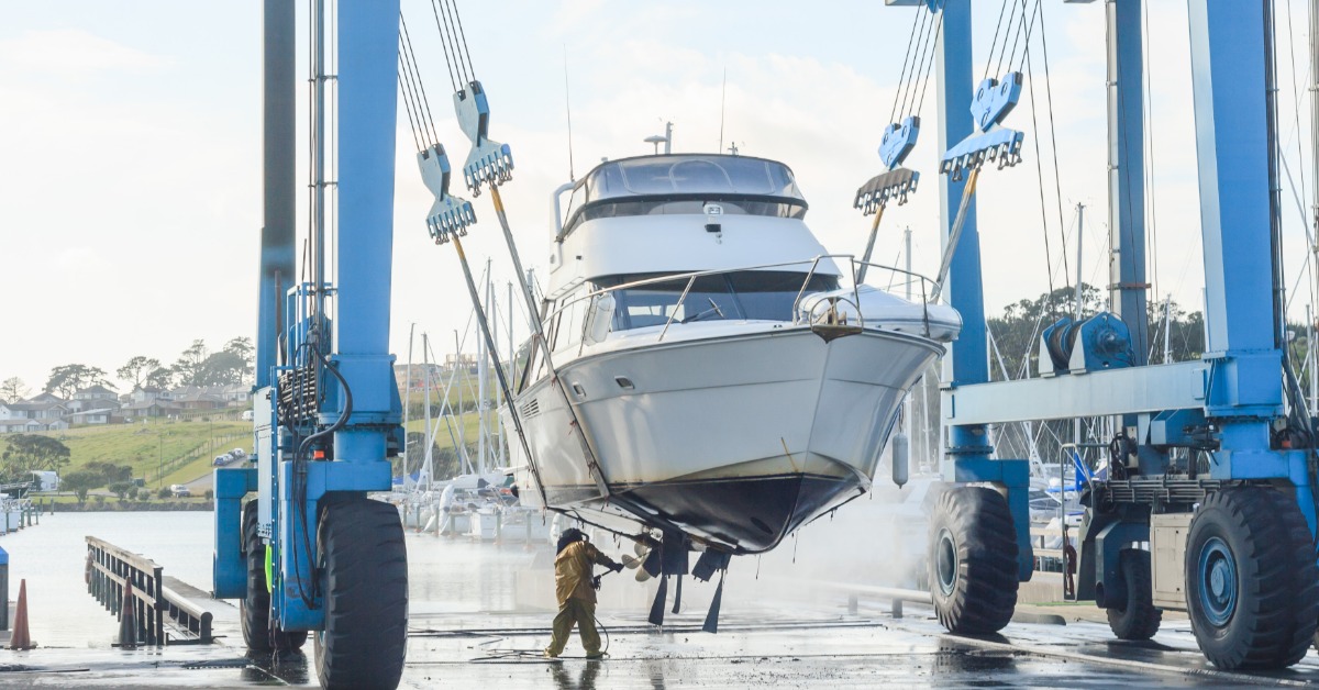 Large white yacht hangs from a blue boat lift above a marina work area as a worker stands beneath the raised vessel.
