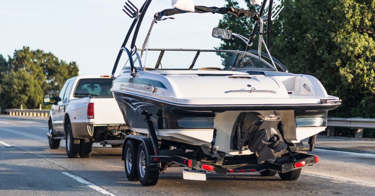 White and black motorboat with wakeboard tower secured to a dual-axle trailer, towed by a white pickup truck on a highway.