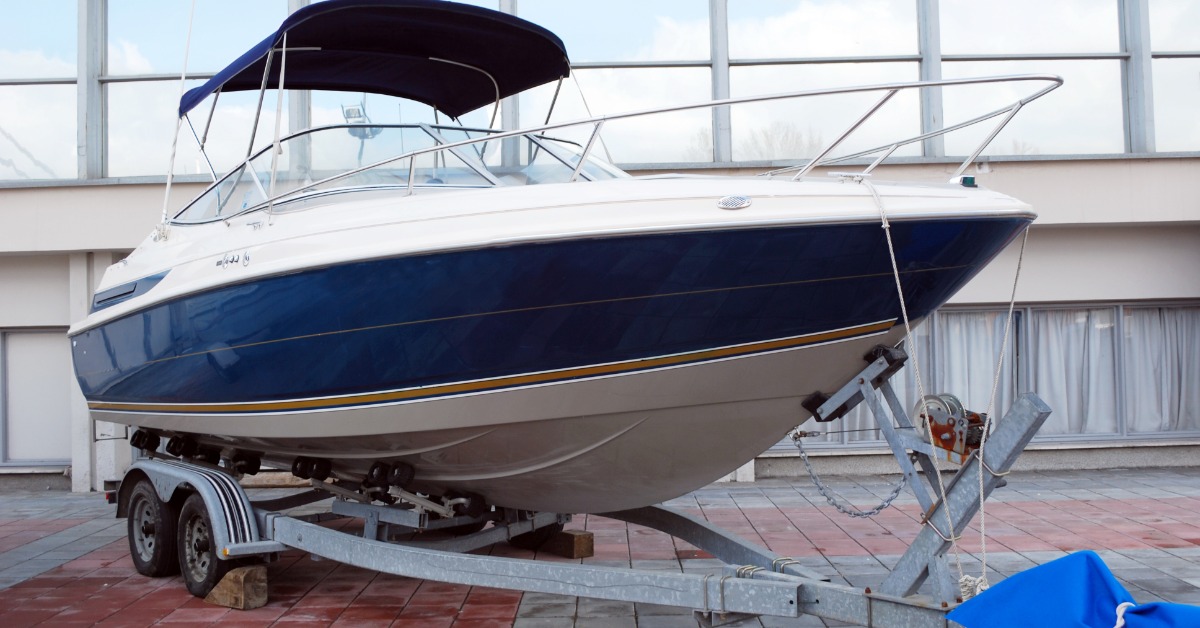 Blue and white motorboat with a navy canopy resting on a dual-axle metal trailer outside a building with large windows.