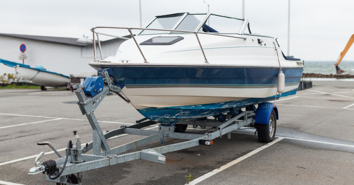 Blue and white cabin motorboat with metal railings resting on a single-axle trailer in a waterfront parking lot.